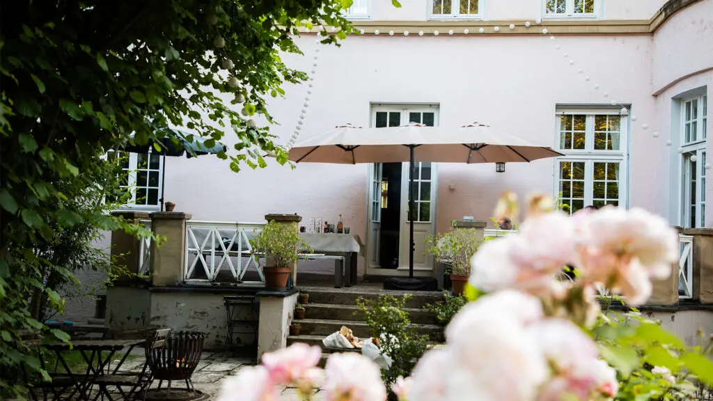 Ein gemütlicher Hinterhof mit einem blassrosa Haus, Sonnenschirmen und einem Tisch mit Getränken. Blumen und Grünpflanzen umgeben den Sitzbereich und schaffen eine entspannte Atmosphäre. Darüber hängen Lichterketten.