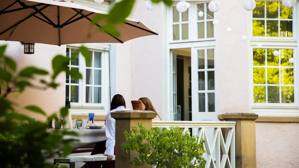 Zwei Personen sitzen an einem Tisch im Freien unter einem großen Sonnenschirm auf einer Terrasse, die von Grün umgeben ist. Darüber hängen weiße Lichterkugeln, und das Gebäude hinter ihnen hat große Fenster und cremefarbene Wände.