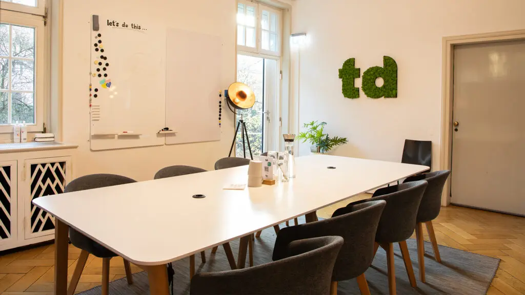 Ein moderner Besprechungsraum mit einem langen weißen Tisch, acht dunklen Stühlen, einem Whiteboard, einer Lampe und einer Wanddekoration mit grünen Moosbuchstaben td. Große Fenster sorgen für natürliches Licht.