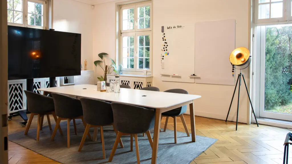Ein moderner Besprechungsraum mit einem weißen Tisch, sechs dunklen Stühlen, einem großen Fernseher auf einem Ständer, einem Whiteboard, einer hohen Stehlampe, Topfpflanzen und großen Fenstern, die natürliches Licht hereinlassen.
