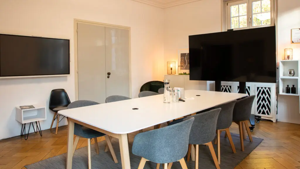 Moderner Konferenzraum mit weißem Tisch, sechs grauen Stühlen, zwei großen Flachbildschirmen, minimalistischem Dekor, Holzboden, kleinen Regalen und Fenstern, die natürliches Licht hereinlassen.