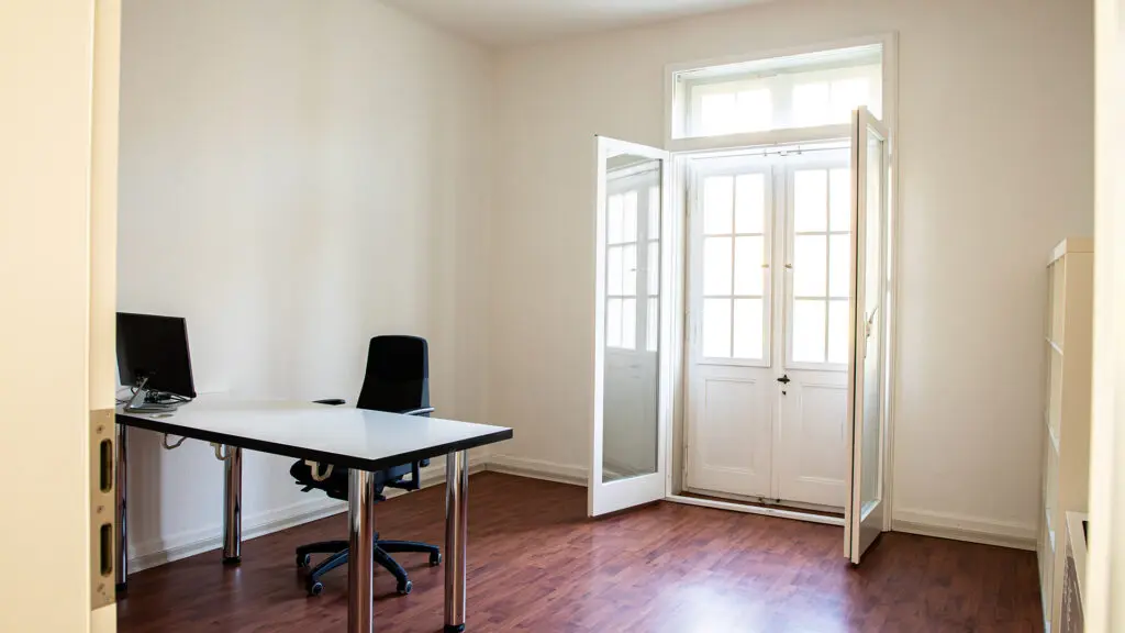 Ein minimalistisches Büro mit einem Schreibtisch, einem schwarzen Stuhl, einem Computermonitor und einer Tastatur. Der Raum hat weiße Wände, einen Holzfußboden und eine Glastür, die natürliches Licht hereinlässt.