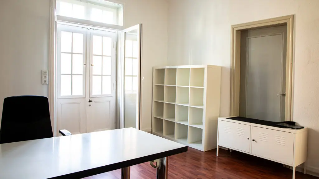 Ein helles, minimalistisches Büro mit einem weißen Schreibtisch, einem schwarzen Stuhl, einem leeren weißen Regal und einem kleinen Schrank. Das Sonnenlicht fällt durch Glastüren und Fenster und beleuchtet den Holzboden.