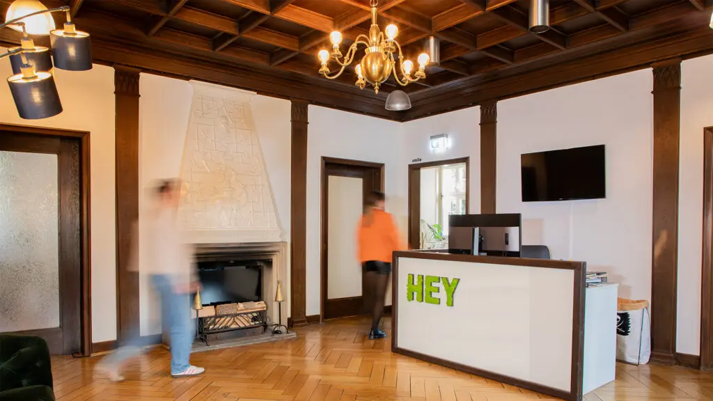 Eine moderne Bürolobby mit Holzböden und -decken, einem weißen Empfangstresen mit der Aufschrift HEY, einem Fernseher, einem Kamin und zwei Personen, die scheinbar unscharf in Bewegung sind. Warme Beleuchtung und modernes Dekor füllen den Raum.