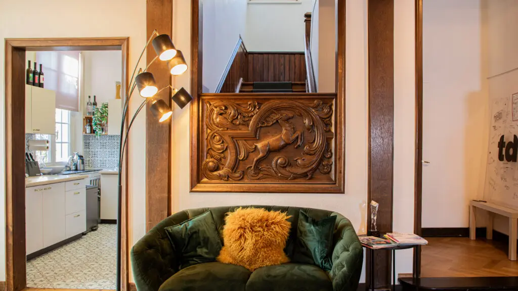 Ein gemütlicher Wohnbereich mit einem grünen Samtsofa und goldenen Kissen, einer modernen Stehlampe, einer verschnörkelten Holzwand und einem Blick in eine Küche im Vintage-Stil und eine Treppe mit dunklen Holzdetails.
