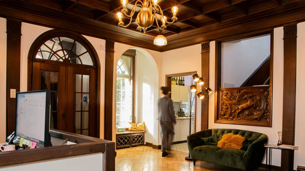 Eleganter Büroraum mit Holzakzenten, einem Kronleuchter, einem grünen Samtsofa, großer geschnitzter Wandkunst und einer Person im Anzug, die auf eine helle gewölbte Türöffnung zugeht. Im Vordergrund ist ein Schreibtisch mit einem Computer zu sehen.