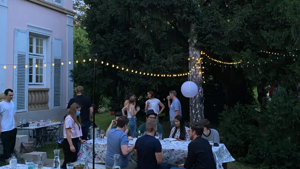 Menschen versammeln sich in einem Garten zu einer Abendgesellschaft, sitzen und stehen um Tische mit Speisen und Getränken. Lichterketten und Laternen sind zwischen Bäumen aufgehängt und schaffen eine festliche Atmosphäre in der Nähe eines hellrosa Gebäudes.