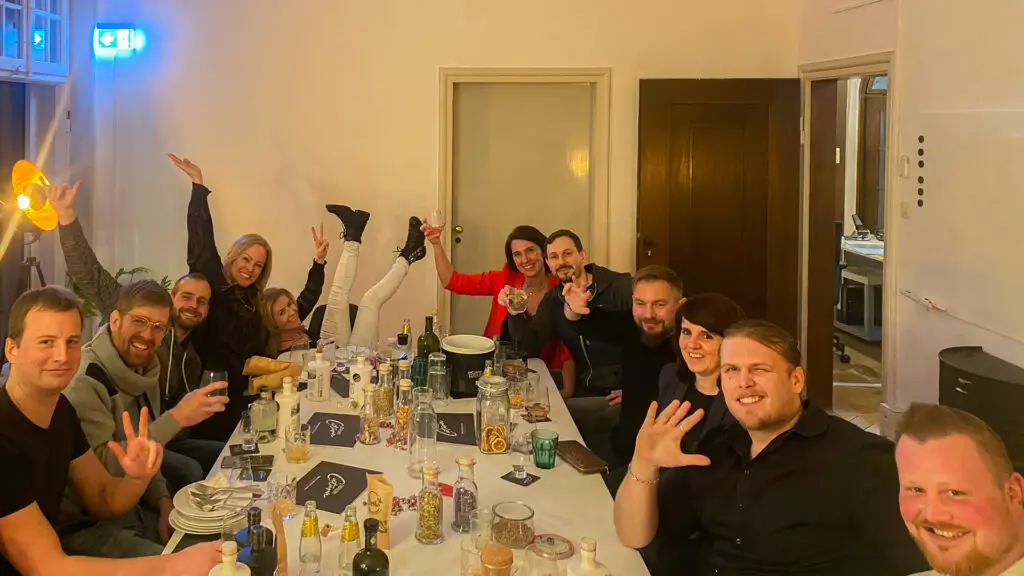 Eine Gruppe von zwölf Personen sitzt und posiert fröhlich um einen mit Essen, Getränken und Kerzen gedeckten Esstisch, einige winken oder machen Friedenszeichen und schaffen so eine lebhafte, feierliche Atmosphäre im Haus.