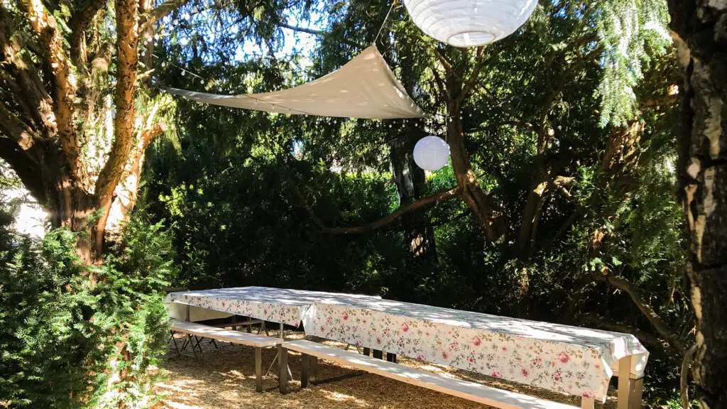 Ein langer Picknicktisch mit geblümten Tischtüchern steht unter einem weißen Sonnensegel und hängenden Papierlaternen in einem von Bäumen umgebenen, sonnendurchfluteten Garten.