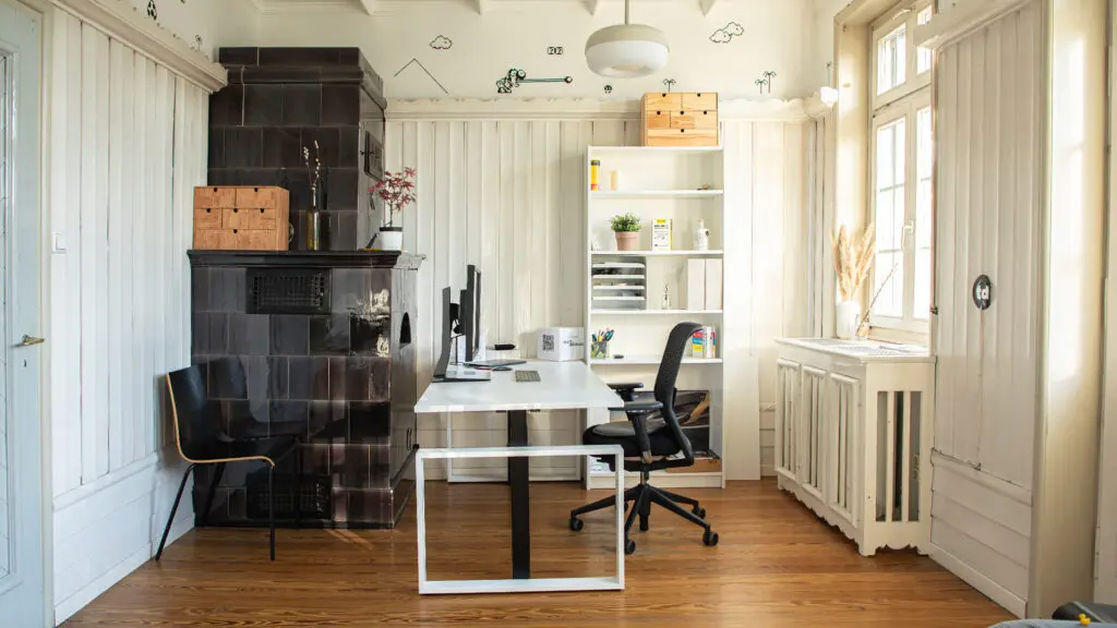 Helles Heimbüro mit weißem Schreibtisch, schwarzem Stuhl, Regalen mit Büchern und Dekoration, schwarzem Kamin, Holzböden und großen Fenstern, die viel Tageslicht hereinlassen.
