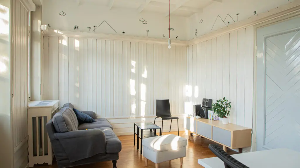 Ein helles, minimalistisches Wohnzimmer mit weiß getäfelten Wänden, Holzboden, einem grauen Sofa, zwei schwarzen Sesseln, einer kleinen Ottomane, einem niedrigen Holzschrank mit Pflanzen und Lautsprechern und skurriler schwarzer Linienkunst an der Decke.