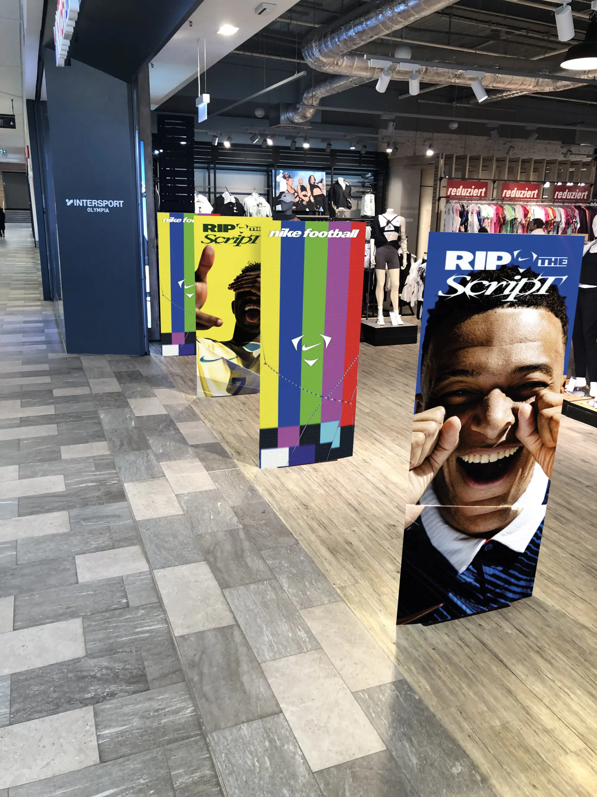Hohe, farbenfrohe Schilder mit lächelnden Sportlern und der Aufschrift „RIP THE SCRIPT“ sowie Nike-Fußballartikel säumen den Eingang eines modernen Sportbekleidungsgeschäfts, im Hintergrund sind Schaufensterpuppen und Kleidung zu sehen.