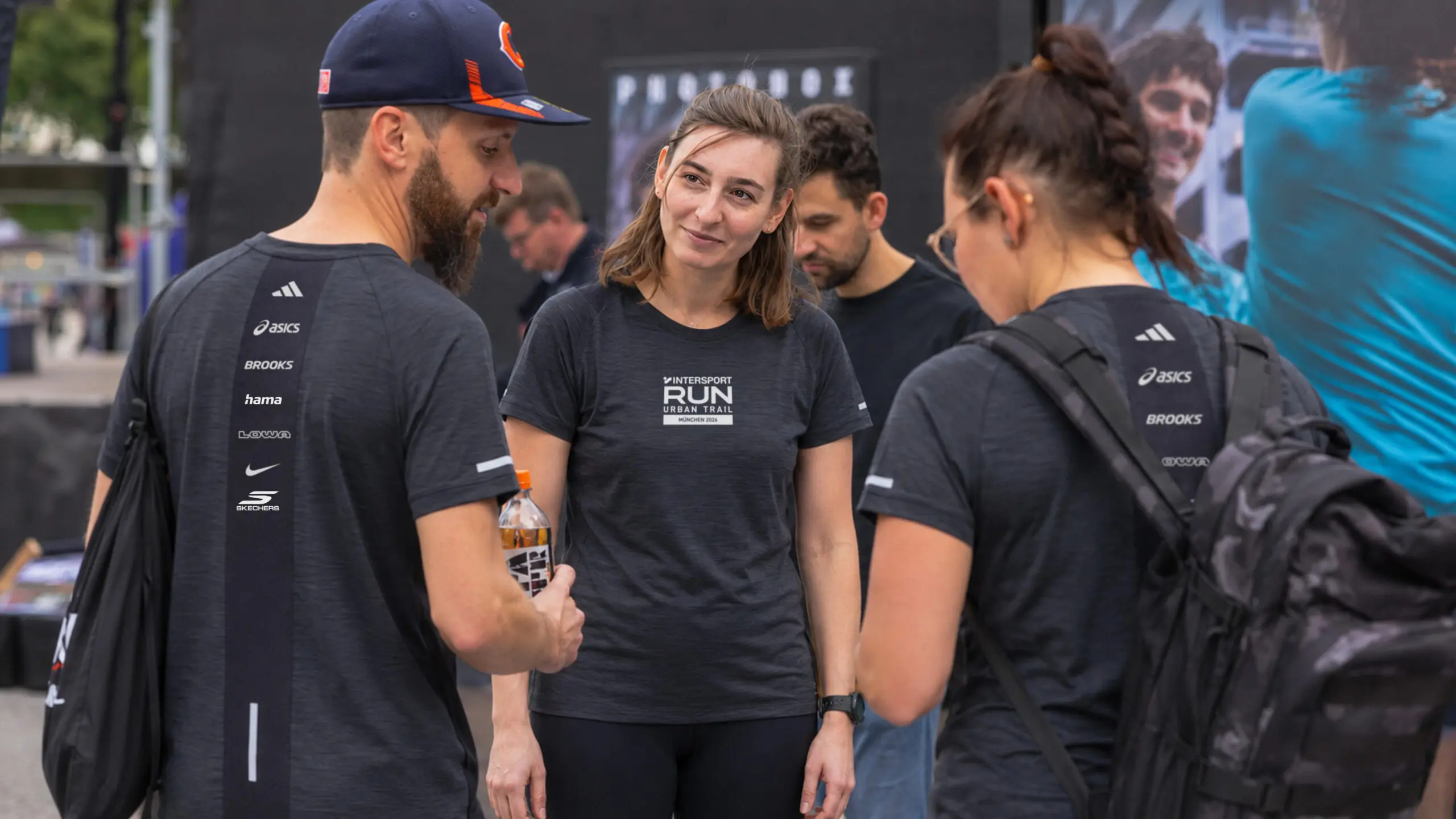 Eine Gruppe von Menschen in einheitlichen Sportshirts steht gemeinsam im Freien, unterhält sich und lächelt. Eine Frau blickt in die Kamera, während andere wegschauen oder zur Seite schauen. Sie scheinen an einer Laufveranstaltung teilzunehmen.