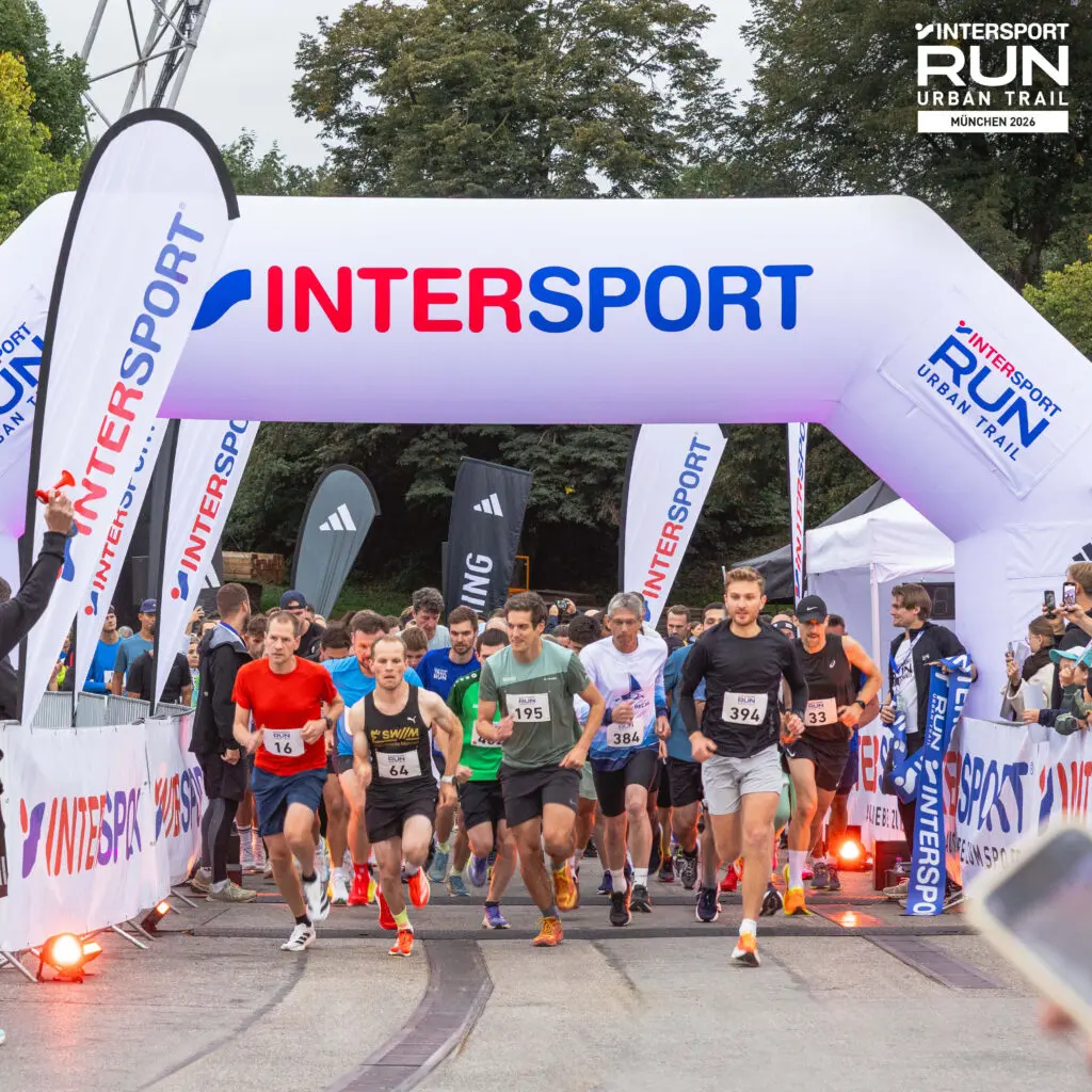 Läufer in Sportbekleidung sprinten bei einem Laufevent im Freien von der Startlinie unter einem weißen, aufblasbaren Torbogen mit der Aufschrift „INTERSPORT“ hindurch, während in der Nähe Zuschauer und Banner zu sehen sind. Bei der Veranstaltung handelt es sich um den „INTERSPORT Run Urban Trail“.