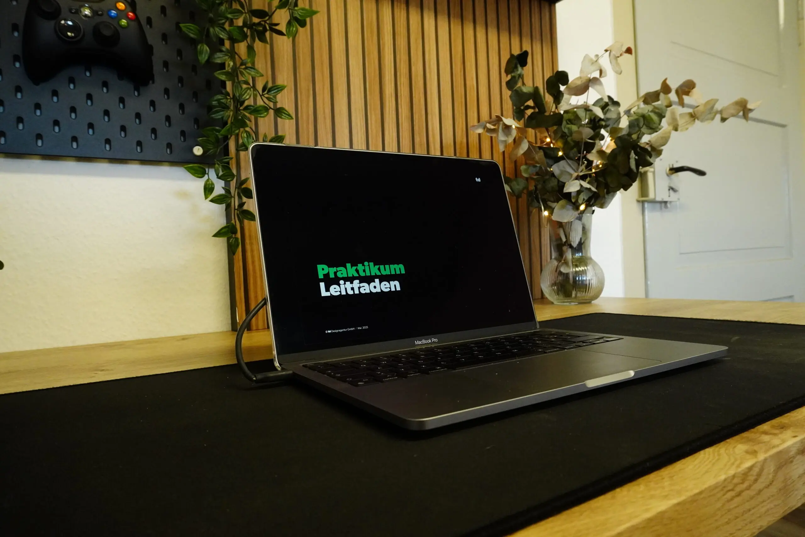 Auf einem Schreibtisch steht ein Laptop, auf dessen Bildschirm die Worte „Praktikum Leitfaden“ zu sehen sind. Auf dem Schreibtisch stehen Pflanzen und Dekoartikel, im Hintergrund ist ein an der Wand befestigter Spiel-Controller zu sehen.