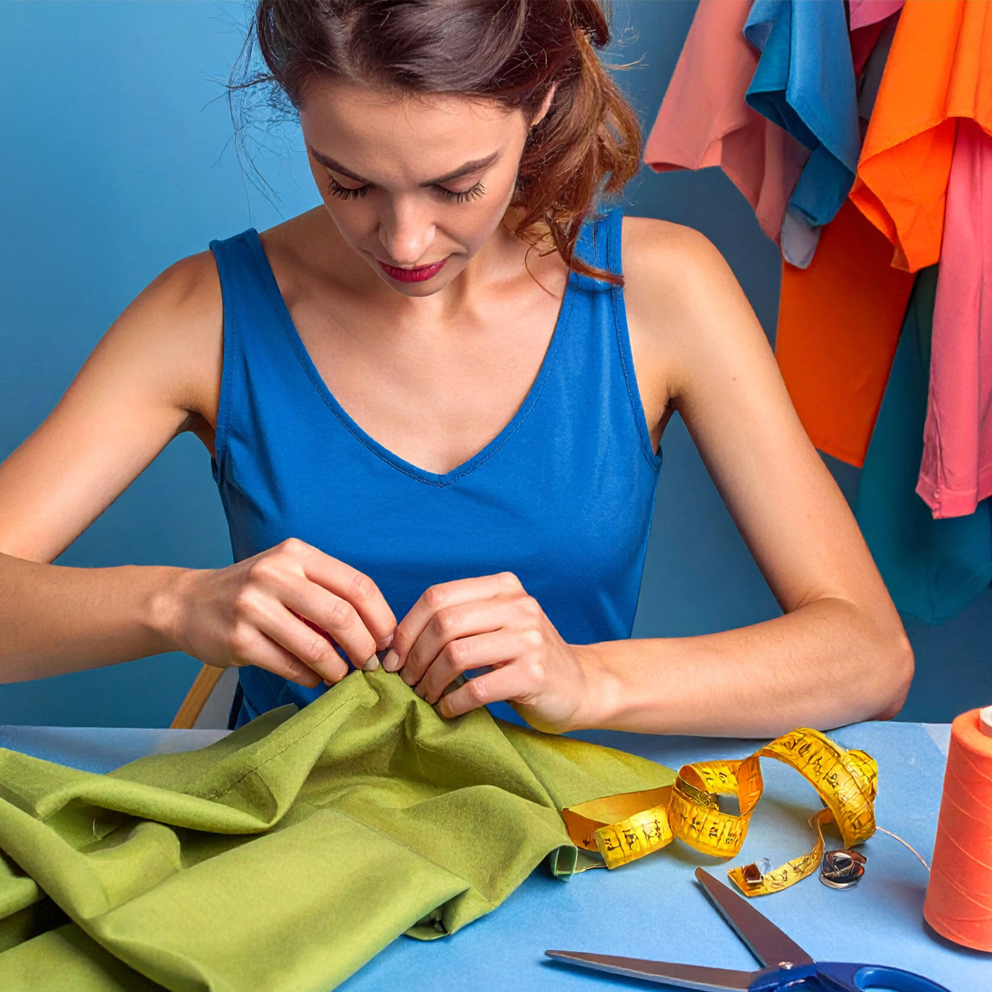 Eine Frau in einem blauen Hemd näht grünen Stoff an einem Tisch, auf dem ein gelbes Maßband, orangefarbener Faden und eine Schere liegen. Im Hintergrund hängen bunte Kleidungsstücke vor einer blauen Wand.