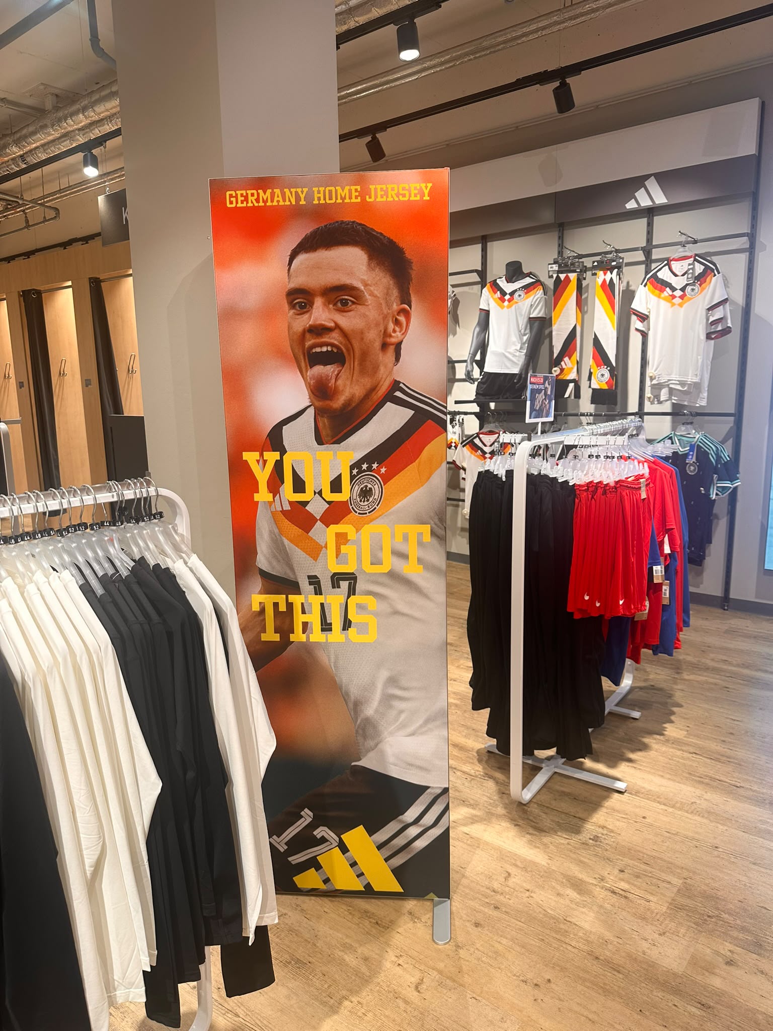 In einem Bekleidungsgeschäft hängt ein großes Plakat, auf dem ein Fußballspieler im deutschen Heimtrikot zu sehen ist, mit der Aufschrift „YOU GOT THIS“. Im Hintergrund sind Kleiderständer mit T-Shirts und deutschen Trikots zu sehen.