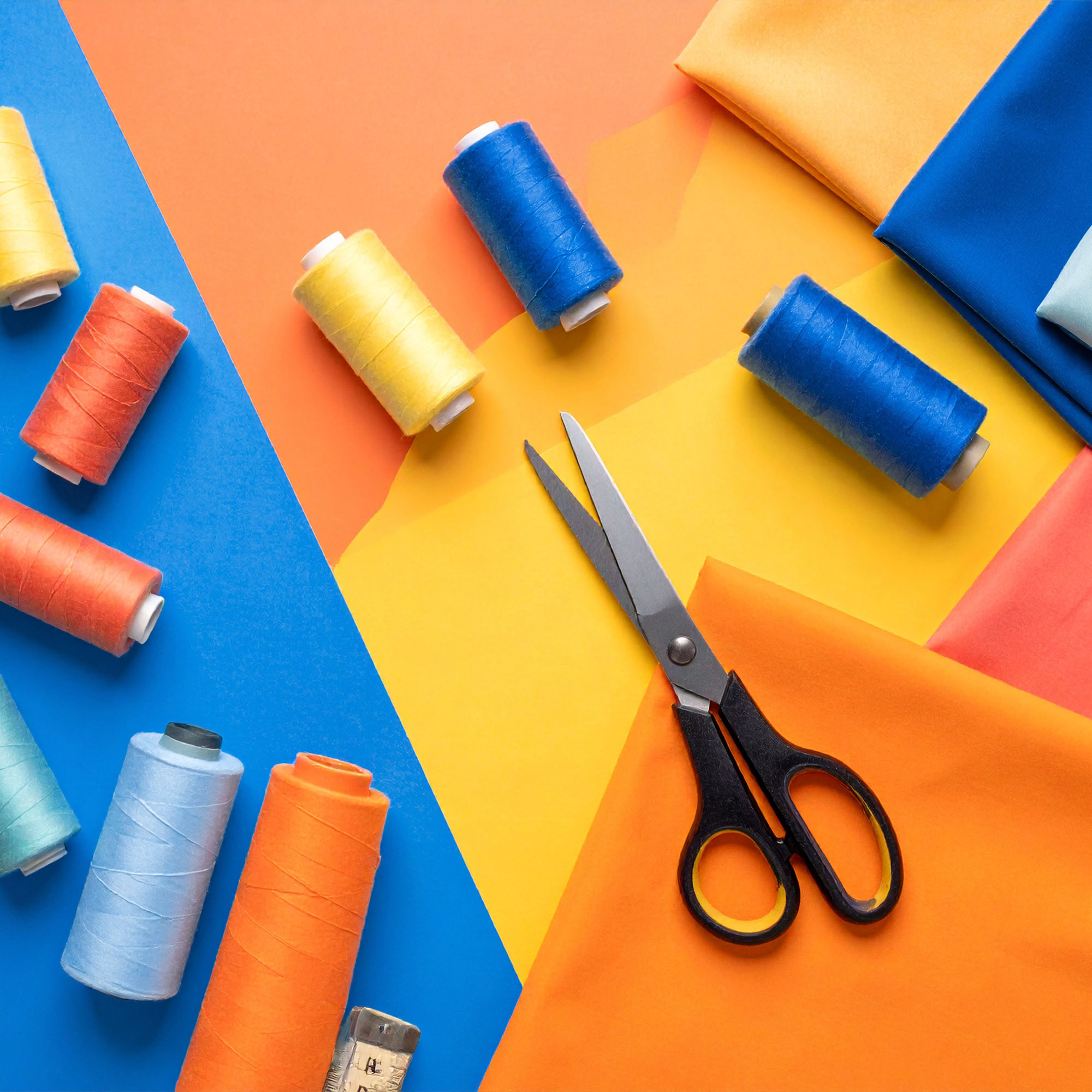 Auf einem blau-orangefarbenen Hintergrund sind Garnrollen in Blau, Gelb und Orange, Stoffbahnen in passenden Farben sowie eine Schere mit schwarzem Griff angeordnet.