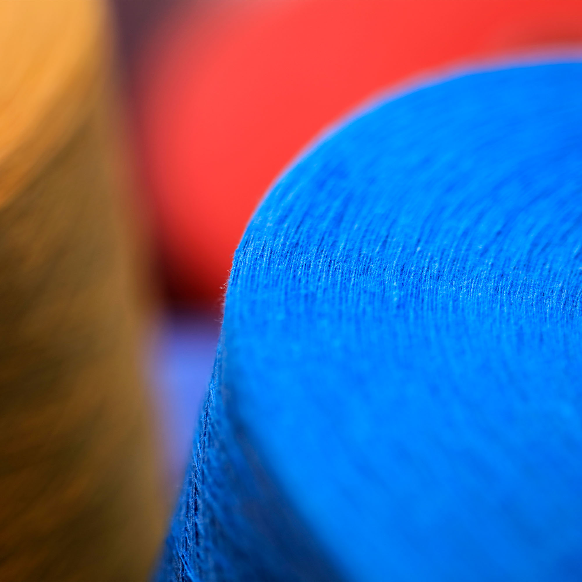 Nahaufnahme eines blauen, zylindrischen Objekts mit strukturierter Oberfläche; im Hintergrund sind unscharfe orangefarbene und rote Objekte zu sehen, die eine abstrakte Komposition bilden.