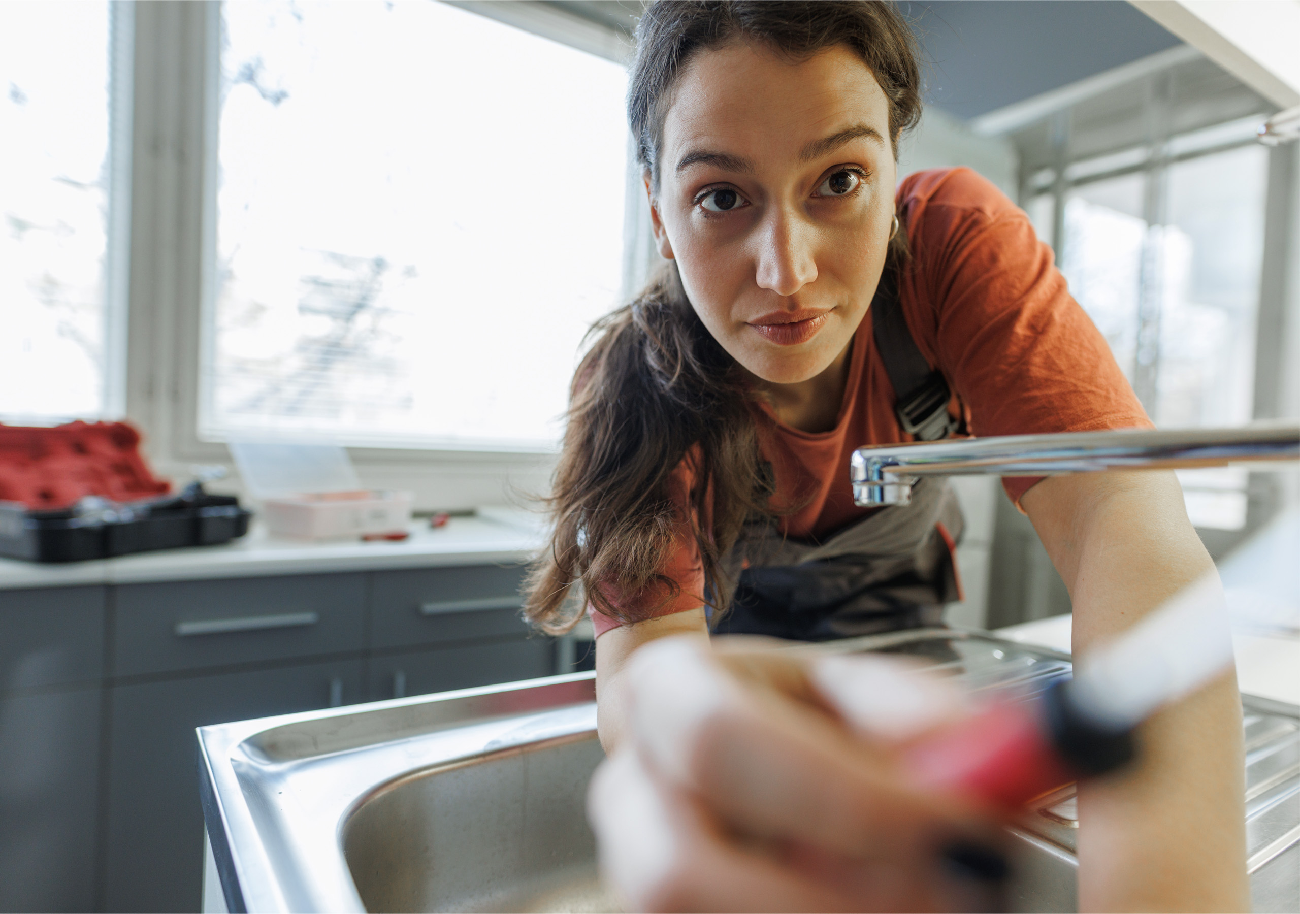 Eine Frau in Arbeitskleidung repariert mit einem Schraubenzieher einen Wasserhahn am Spülbecken, wobei sie sich nach vorne beugt und sich ganz auf ihre Arbeit konzentriert. Im Hintergrund fällt Tageslicht durch die Fenster.